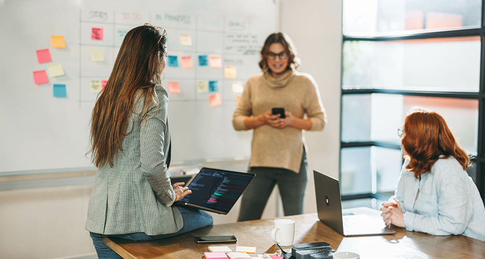 Two women standing talking in a meeting room with a white board in the background
