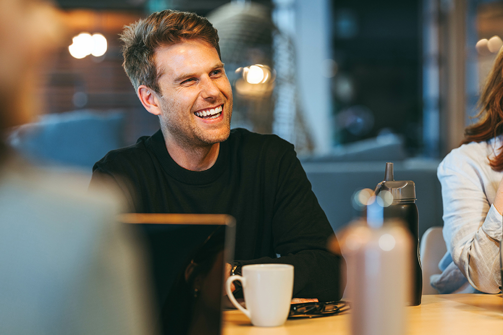 Man smiling sitting at a desk with a coffee cup in front of him