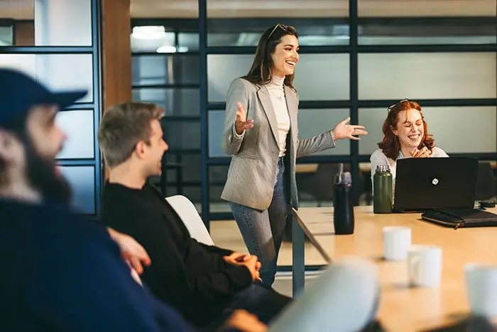 Woman standing at meeting table and speaking, other people at the table are smiling